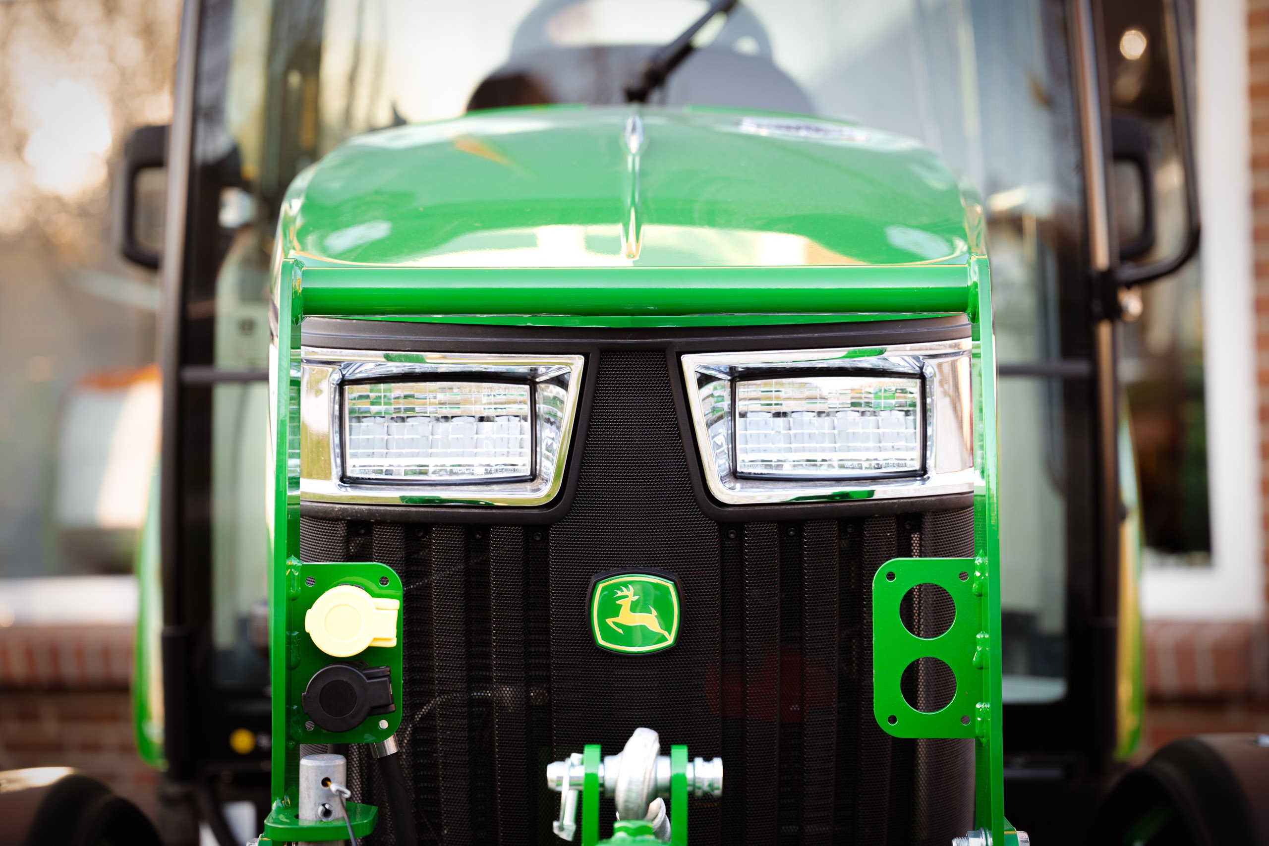 Nahaufnahme der Front eines grünen John Deere Traktors mit Kabine, inklusive Frontkraftheber, elektrischen Anschlüssen und dem markanten springenden Hirsch-Logo im Kühlergrill.