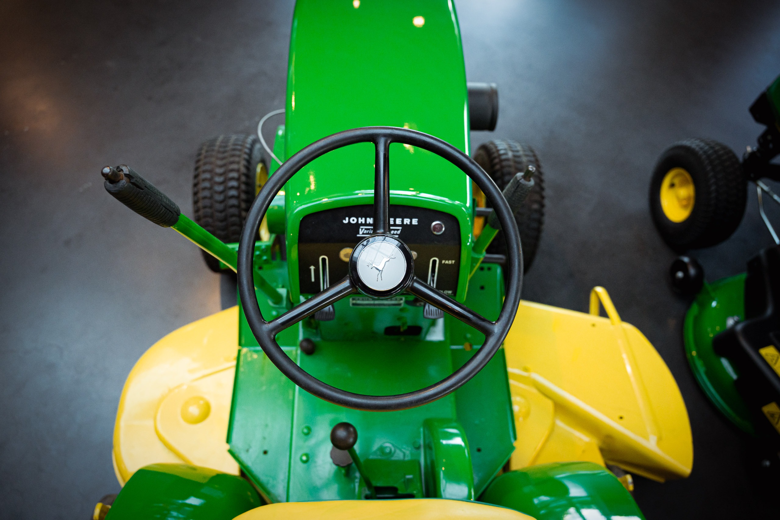 Draufsicht auf das schwarze Dreispeichen-Lenkrad eines grünen John Deere Traktors mit dem springenden Hirsch-Logo im Zentrum und den seitlichen Steuerhebeln.