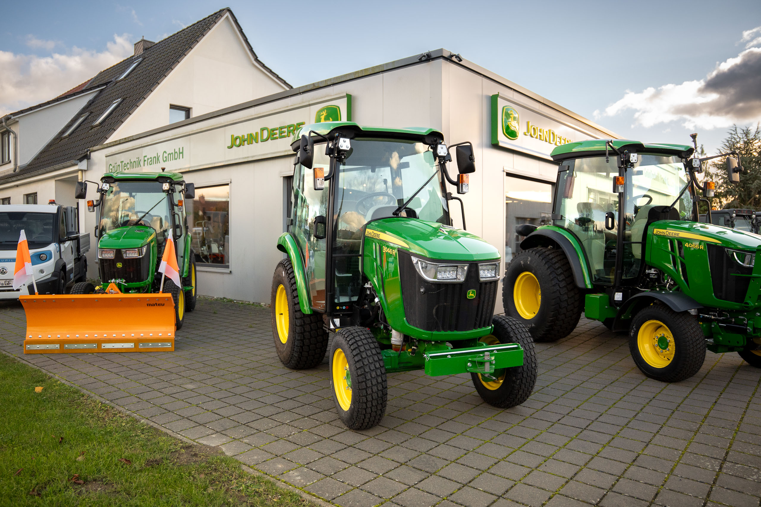 Außenansicht der GrünTechnik Frank GmbH in Neu Roggentin mit drei ausgestellten John Deere Kompakttraktoren, einer davon mit Matev Schneeschild für den Winterdienst.
