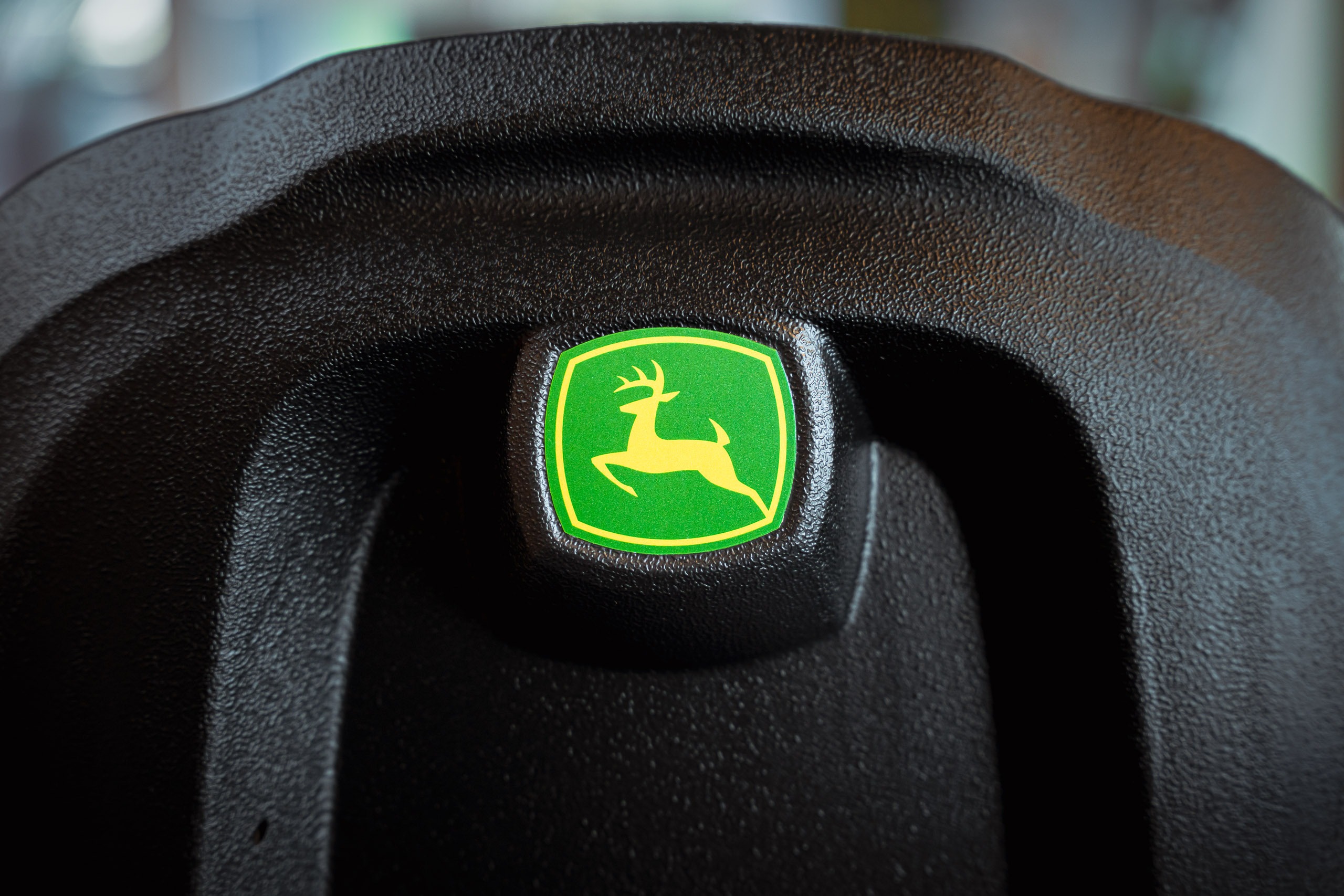 Nahaufnahme des grün-gelben John Deere Logos mit dem springenden Hirsch auf einer strukturierten, schwarzen Kunststoffoberfläche einer Arbeitsmaschine.