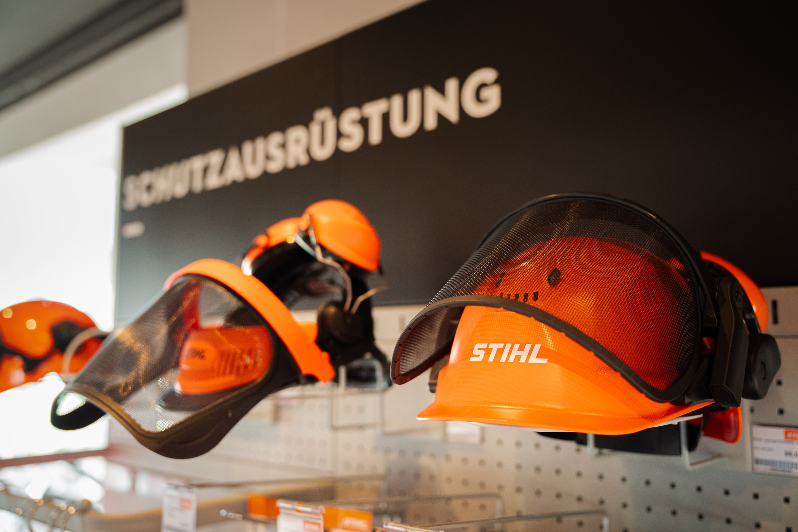 Mehrere orangefarbene STIHL Forsthelme mit integriertem Gehörschutz und Visier, präsentiert an einer Verkaufswand unter dem Schild „SCHUTZAUSRÜSTUNG“.