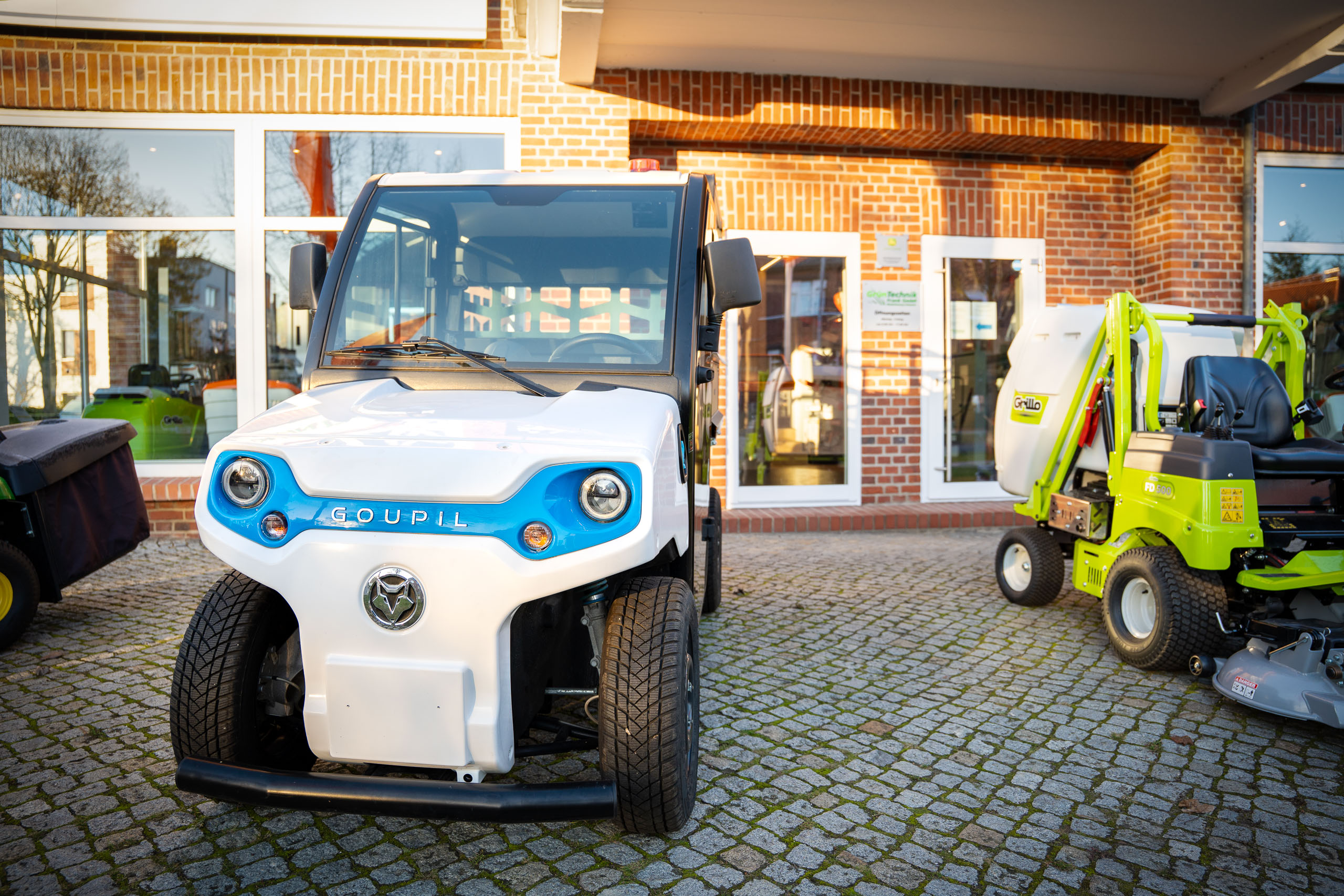 Ein weißes Goupil Elektro-Nutzfahrzeug mit blauen Akzenten parkt auf dem gepflasterten Vorplatz vor dem Backsteingebäude der GrünTechnik Frank GmbH.