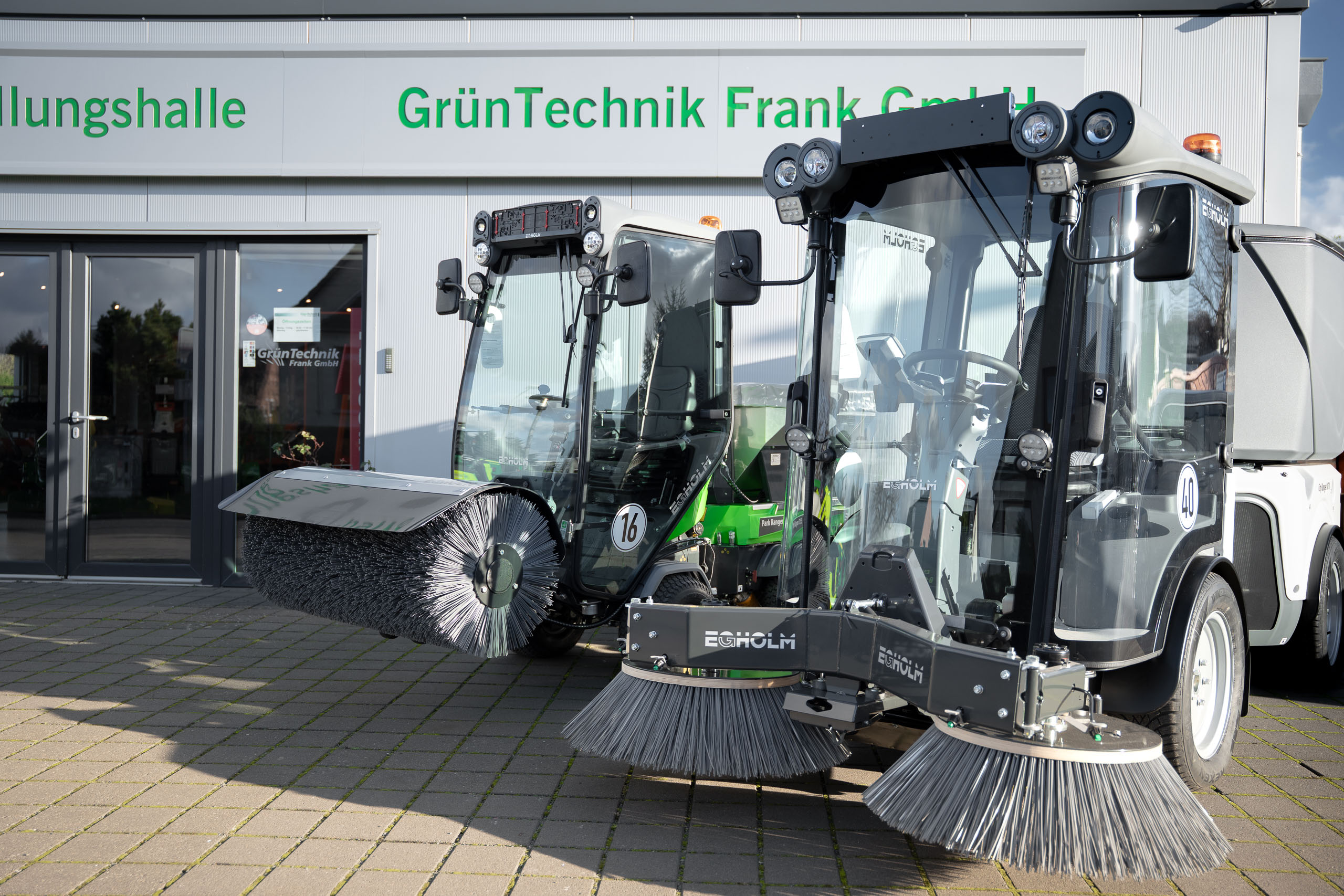 Zwei kompakte Egholm Geräteträger mit unterschiedlichen Kehrbürsten-Aufsätzen für die professionelle Flächenreinigung vor der Ausstellungshalle der GrünTechnik Frank GmbH.
